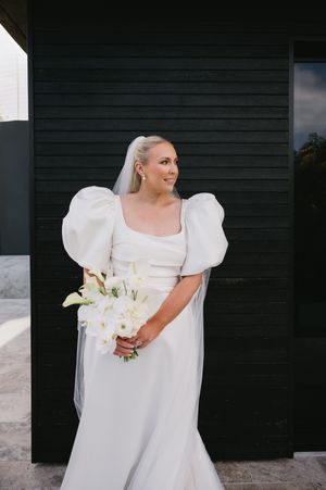 Elegant bridal gown with dramatic puff sleeves and white floral bouquet photographed against a dark exterior wall.