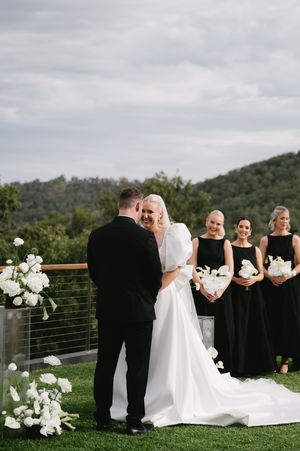 Wedding ceremony setup with black-clad attendants and white floral decorations against mountain backdrop.