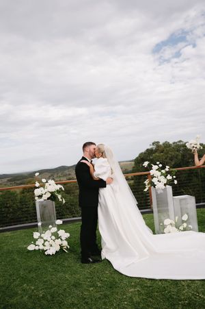 Wedding couple embracing on grassy hilltop with white floral arrangements and mountain backdrop.