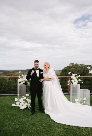 Couple in wedding attire standing on outdoor deck with floral arrangements and mountain view.
