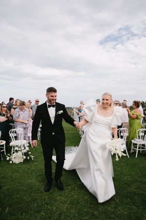 Wedding couple walks across green lawn as guests seated in white chairs applaud.