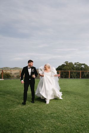 Newlyweds walking together on green lawn with cloudy sky backdrop.
