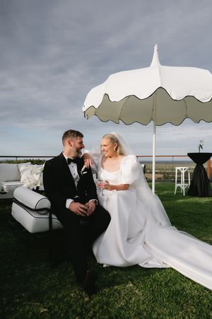 Wedding couple relaxes on white furniture under decorative umbrellas on a lawn.