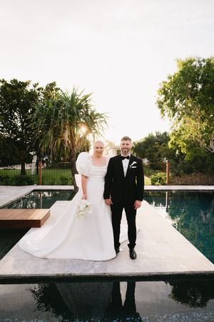Wedding couple walking together on white platform beside infinity pool at sunset.