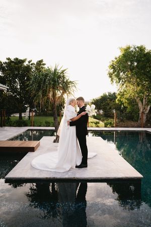 Romantic wedding couple embracing on platform beside infinity pool at sunset.