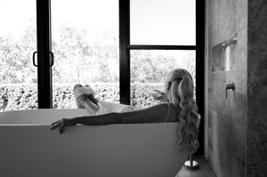 Artistic black and white photo of someone relaxing in a bathtub by a window.