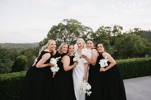 Wedding party in black dresses posing with white bouquets on outdoor terrace.