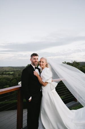 A wedding couple poses on a deck overlooking rolling hills as the bride's veil flows dramatically in the wind.