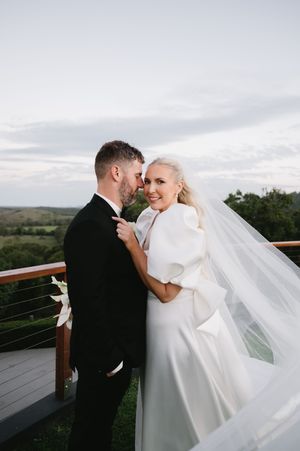 A romantic wedding couple embraces on a deck overlooking rolling hills as the bride's long veil flows in the wind.