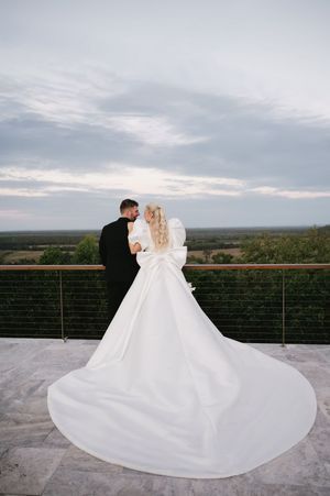 Romantic wedding couple embracing on balcony with dramatic white dress train and scenic view.