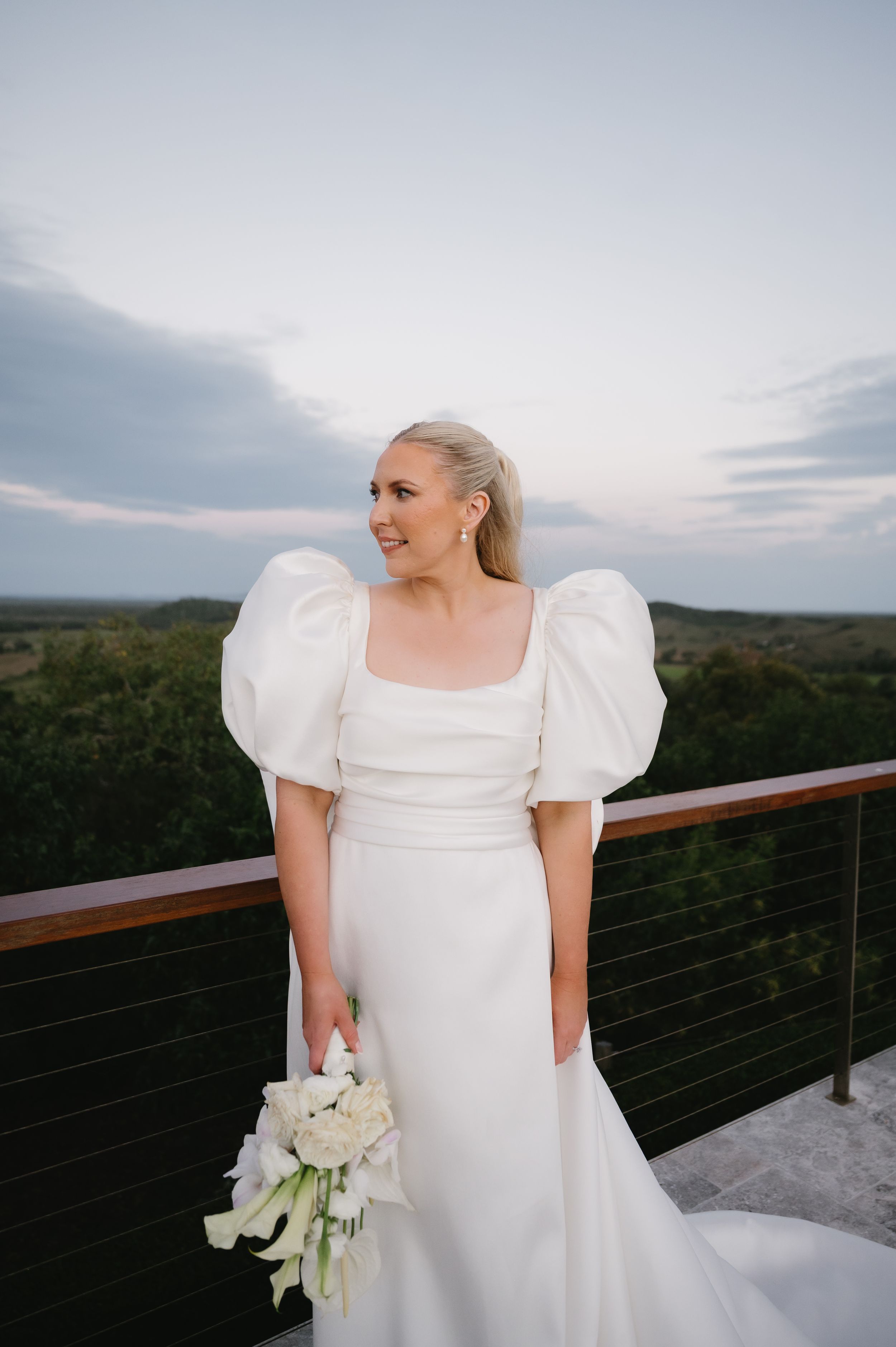 A bride in a classic white wedding dress with puff sleeves poses on a balcony against a scenic mountain landscape at sunset.