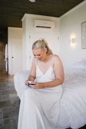 Person in white dress sitting on bed while looking at phone in modern bedroom.