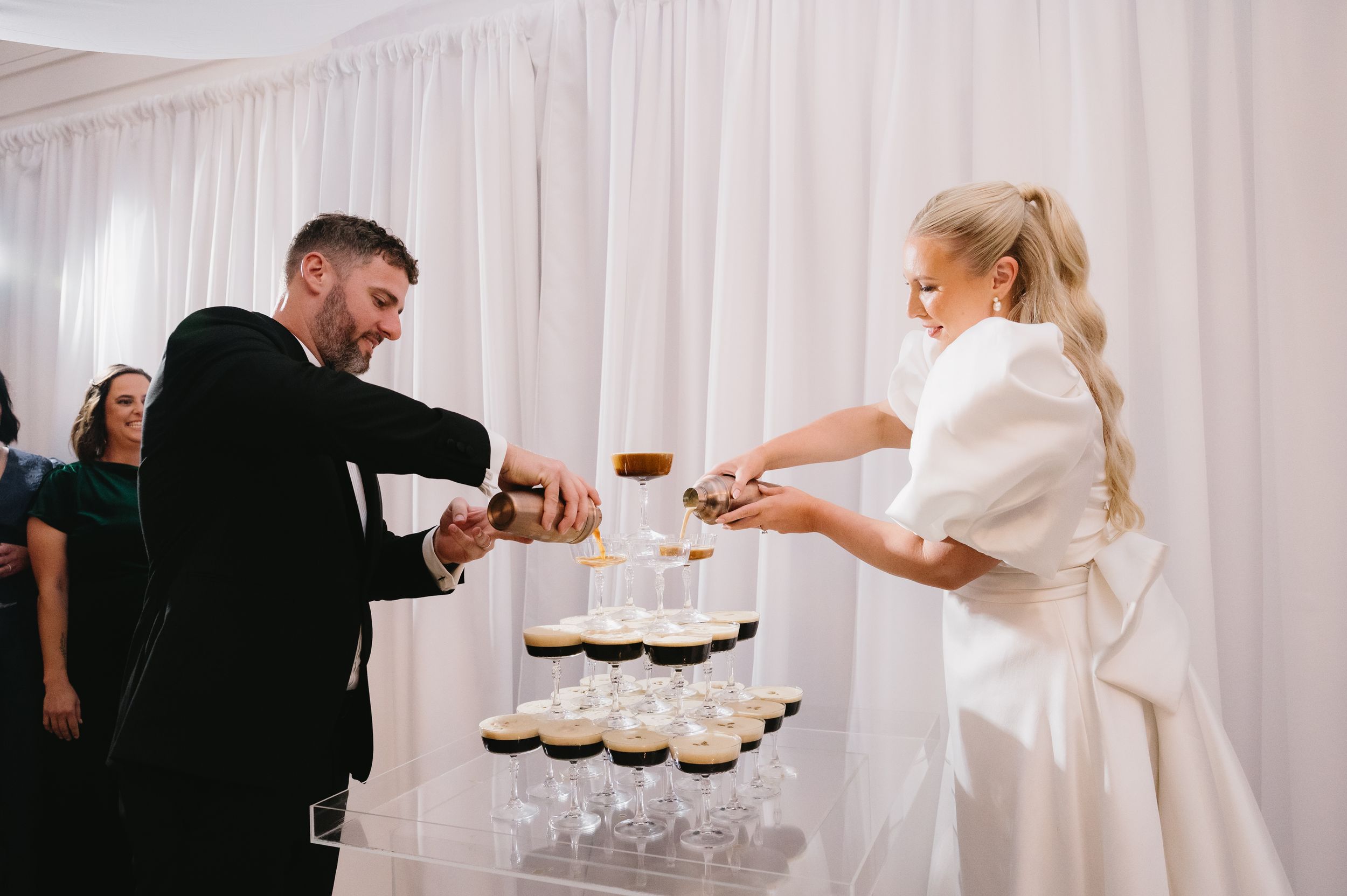 People in formal attire serve drinks on trays at an elegant event with white curtain backdrop.