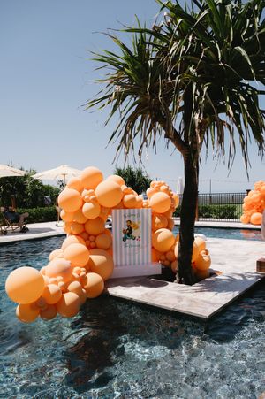 Orange balloon garland floating over a sparkling swimming pool with palm trees in the background.