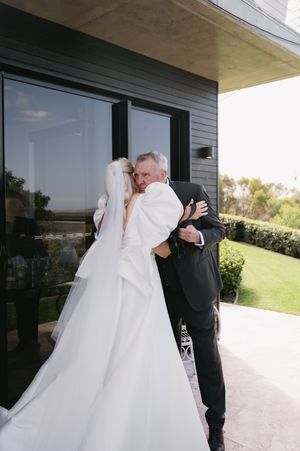 A tender embrace between two people in wedding attire on a patio with glass windows.