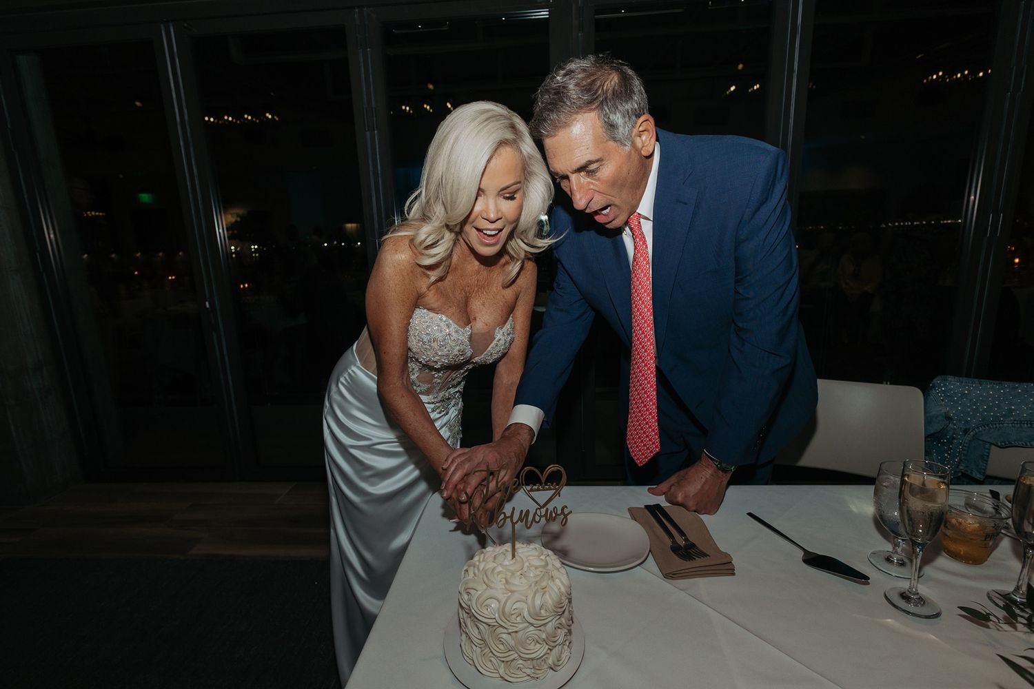A couple in formal attire cuts their white wedding cake together at an evening reception.
