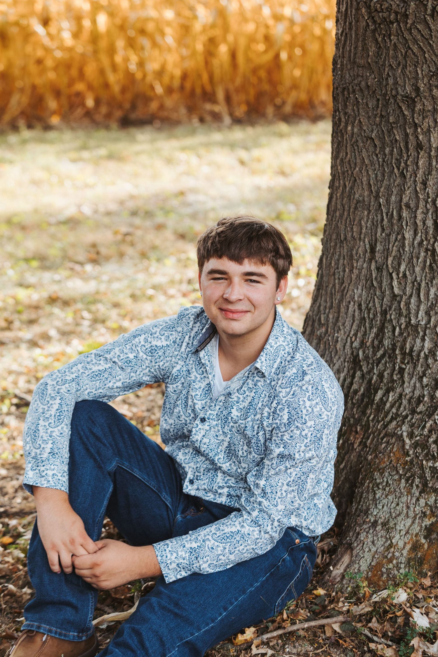 A person wearing a floral print shirt and jeans sits casually against a tree trunk during autumn.