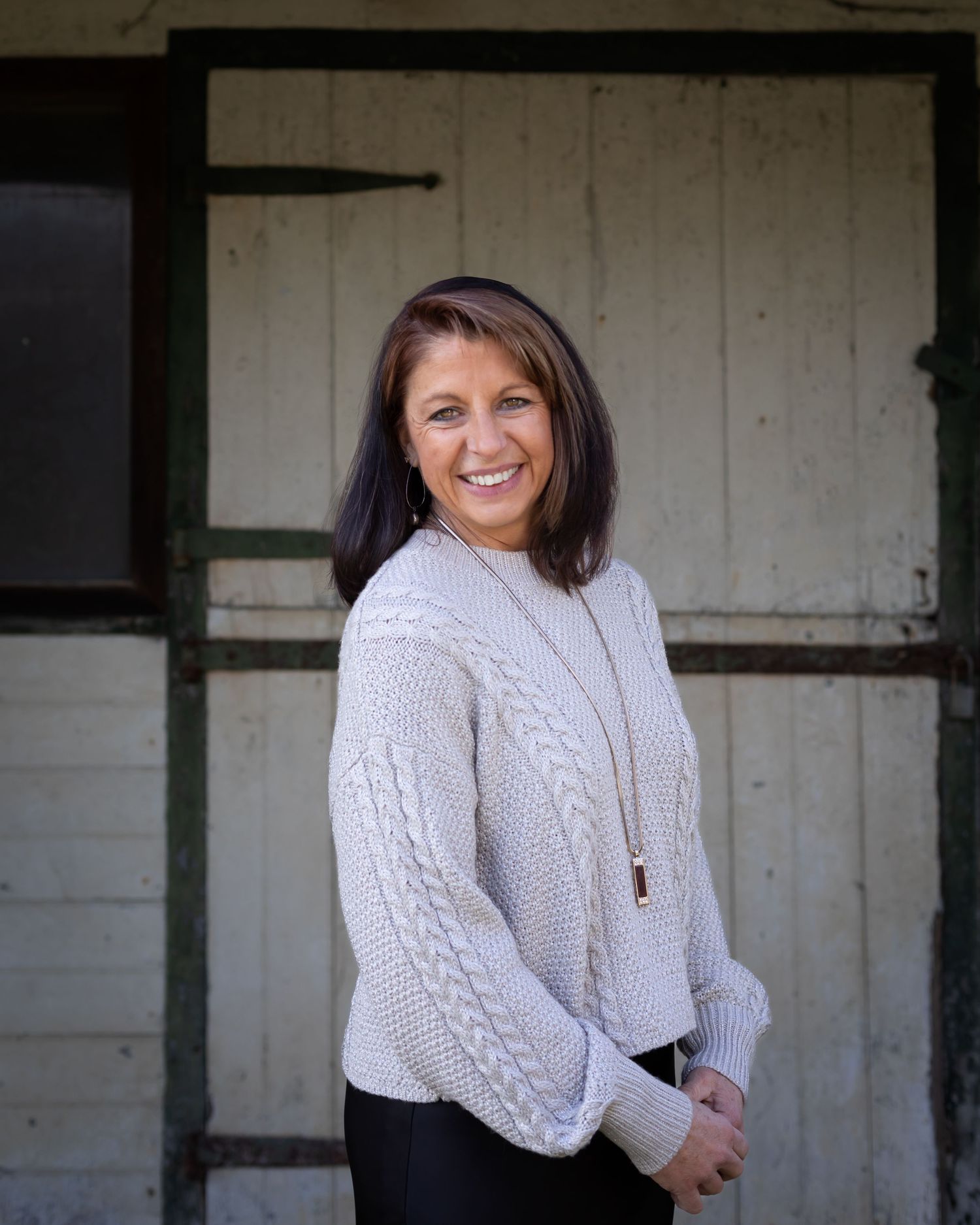 A person wearing a cozy white cable knit sweater and necklace poses outdoors against a rustic white wall.