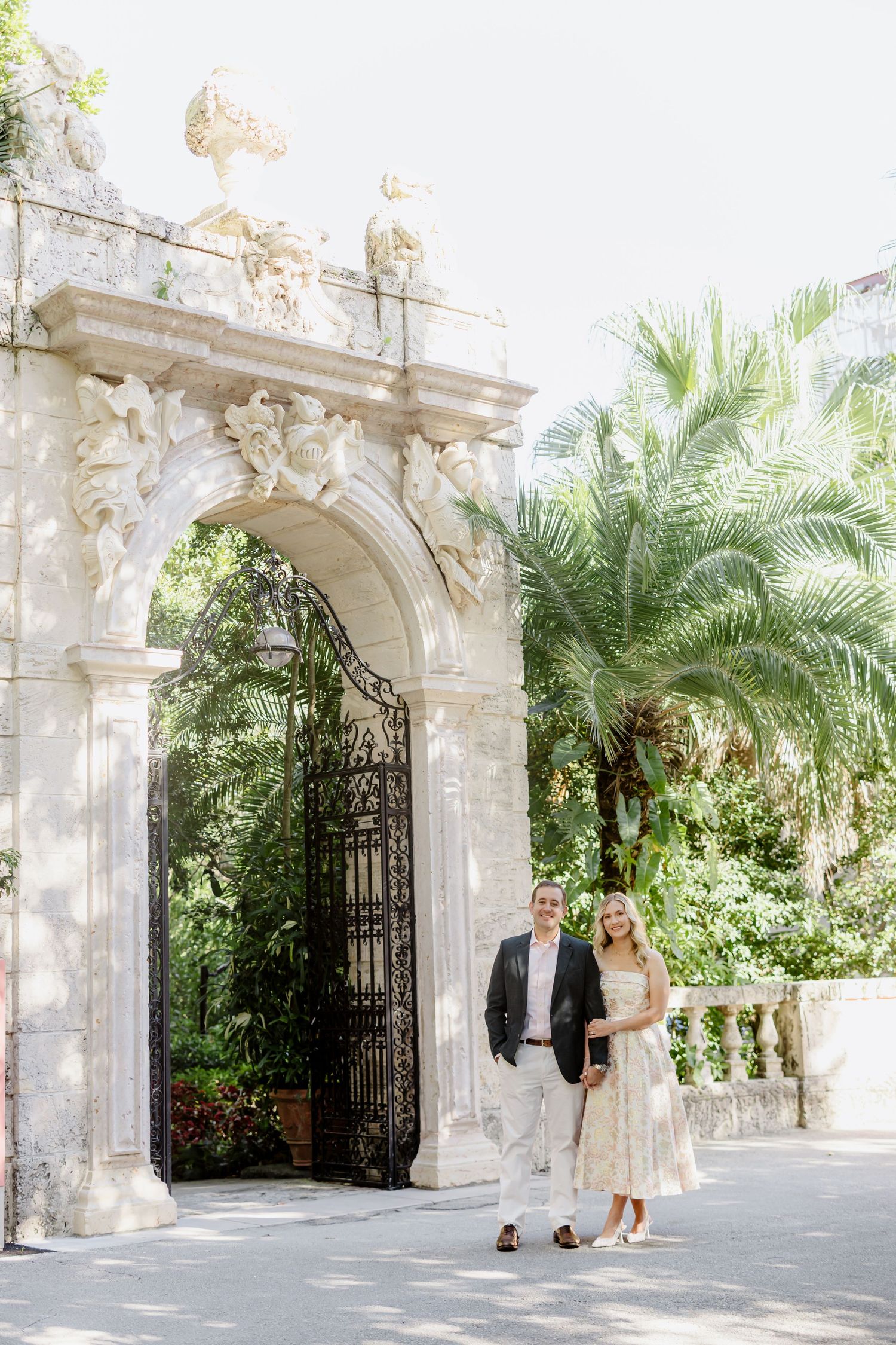 Couple standing beneath an ornate stone archway entrance with palm trees and lush greenery in the background.