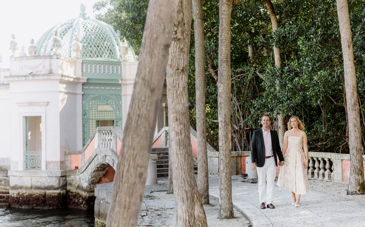 Elegant stone colonnade with ornate architectural details and palm trees in a formal garden setting.