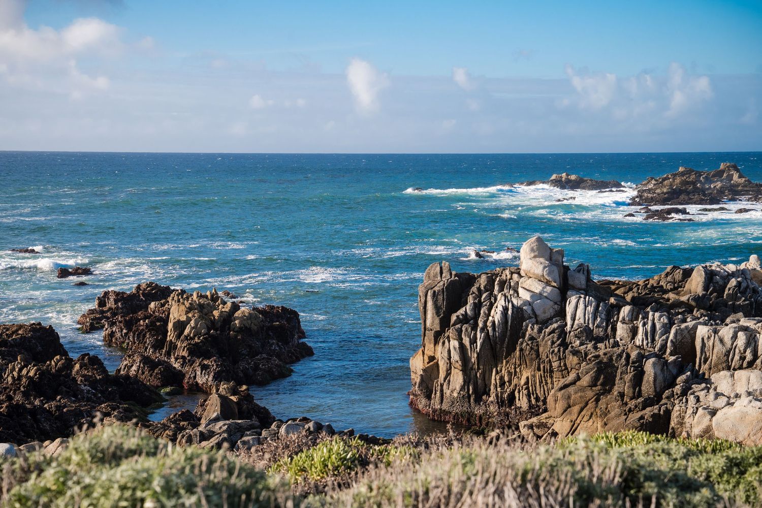 Rocky coastal shoreline with jagged cliffs and crashing waves against a bright blue ocean and sky.