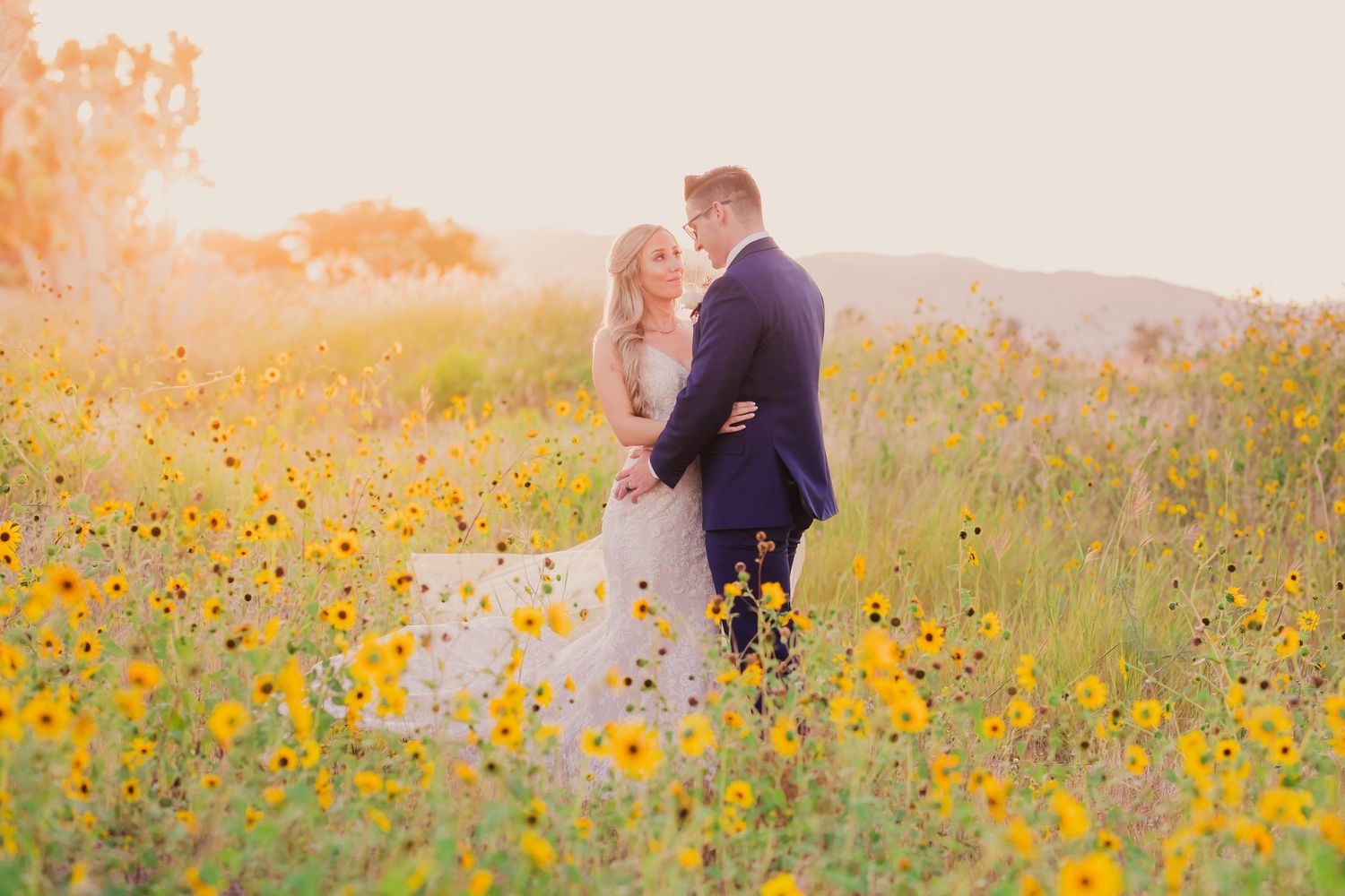 A couple in formal wedding attire walks together through a lush garden path surrounded by trees.