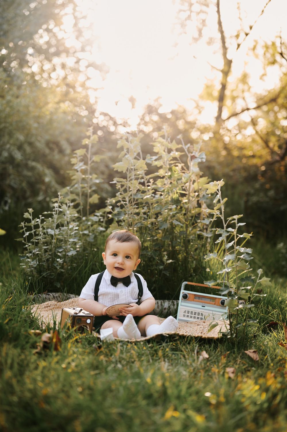 Adorable baby boy in suspenders and bow tie sitting on grass with vintage toy radio outdoors.