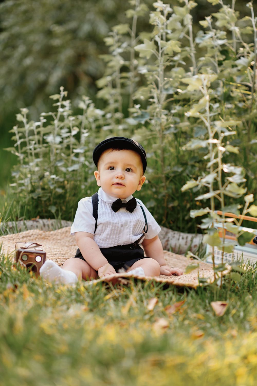 Cute baby boy wearing a black cap, bow tie, and suspenders sitting on a blanket in a garden.