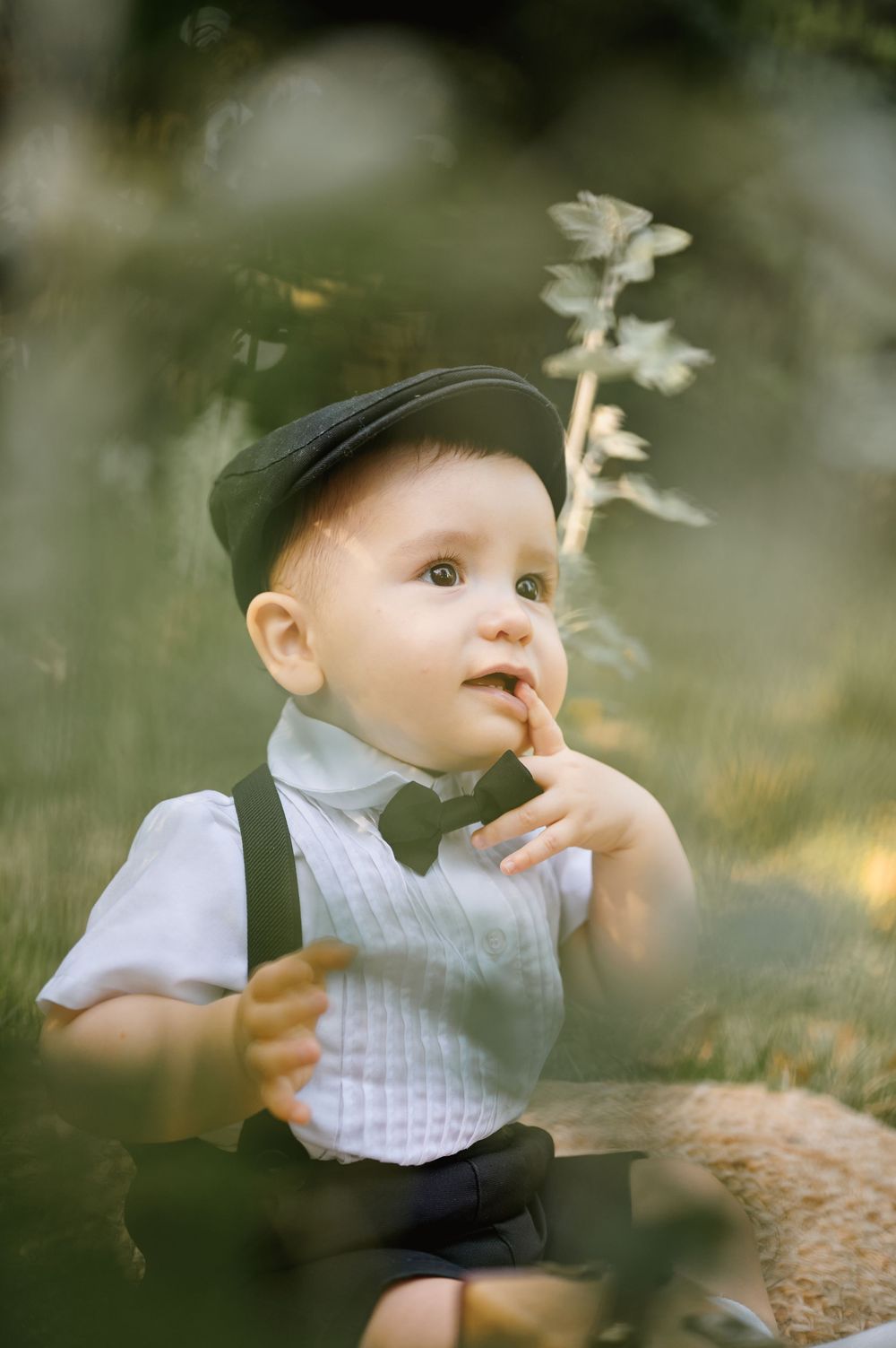Charming baby boy in a flat cap and bow tie sitting outdoors among green plants in soft light.