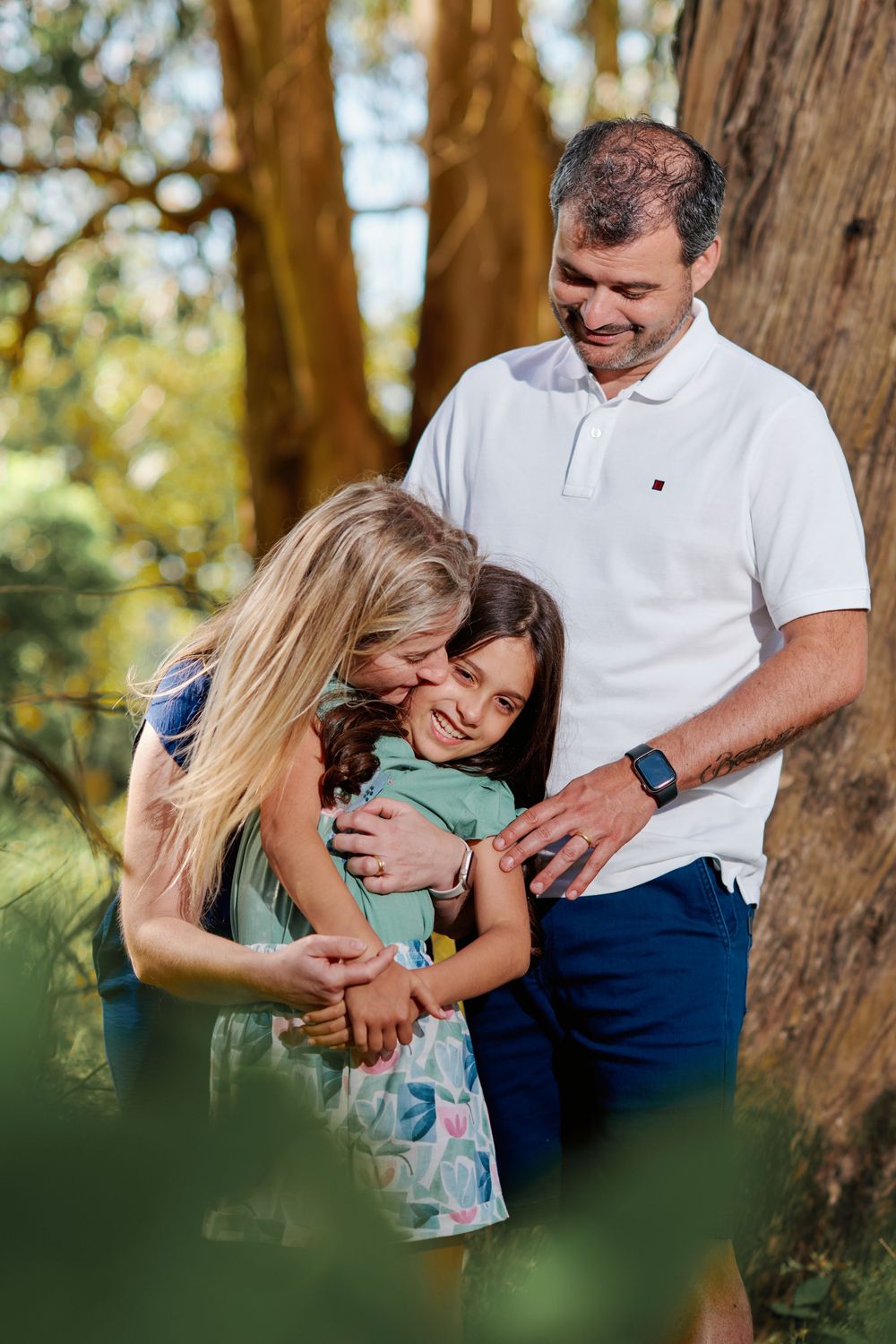 Family of three sharing a joyful hug near a tree in a sunlit forest, mother kissing daughter while father smiles.