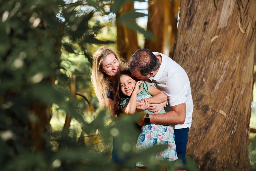 Happy family embracing and laughing together beside a large tree in a sun-dappled forest.