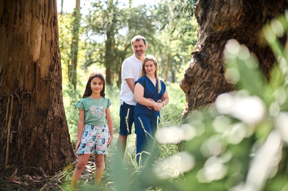 Family of three posing together between large trees in a lush green forest setting.