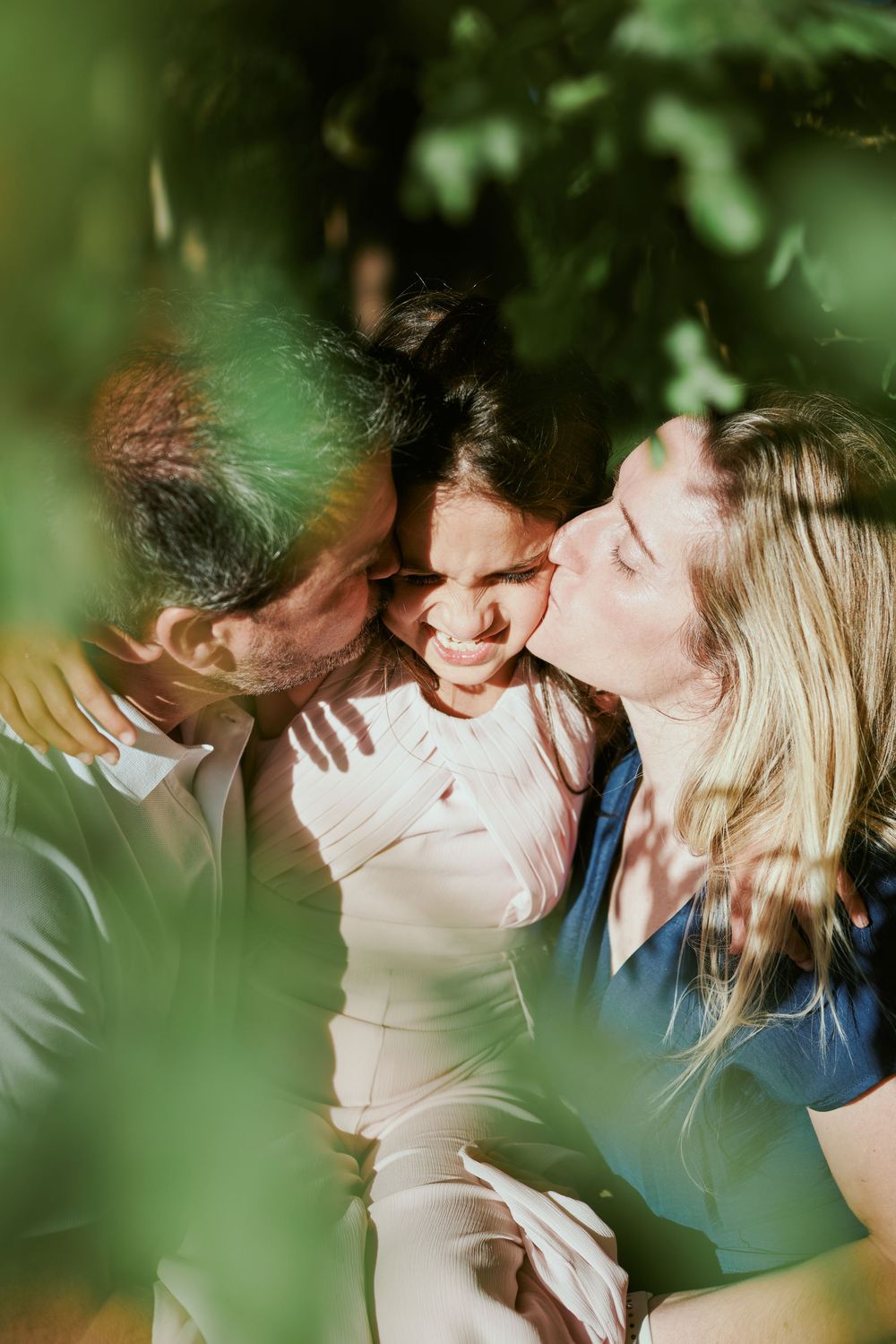 Parents lovingly kissing their child on both cheeks simultaneously, surrounded by lush green foliage.