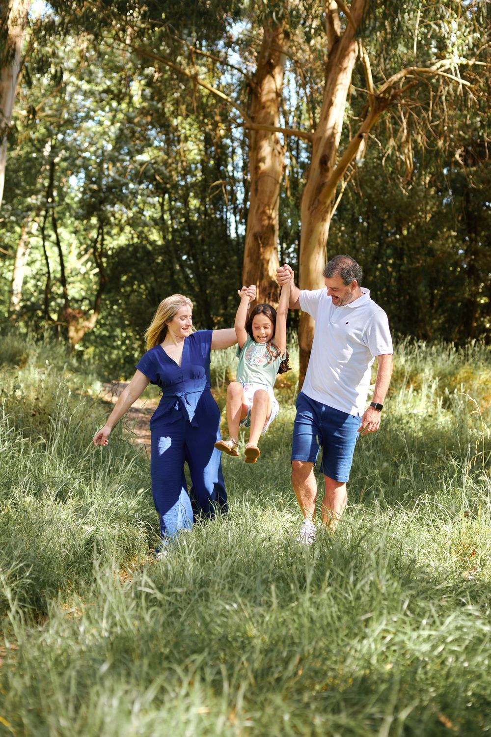 Happy family swinging young daughter between parents in lush green forest clearing.
