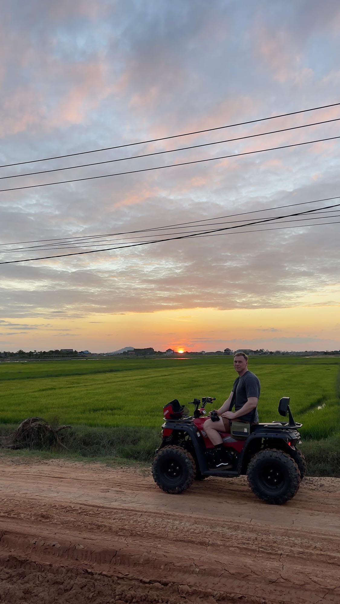 A red ATV drives along a dirt road at sunset with power lines and green fields in the background.