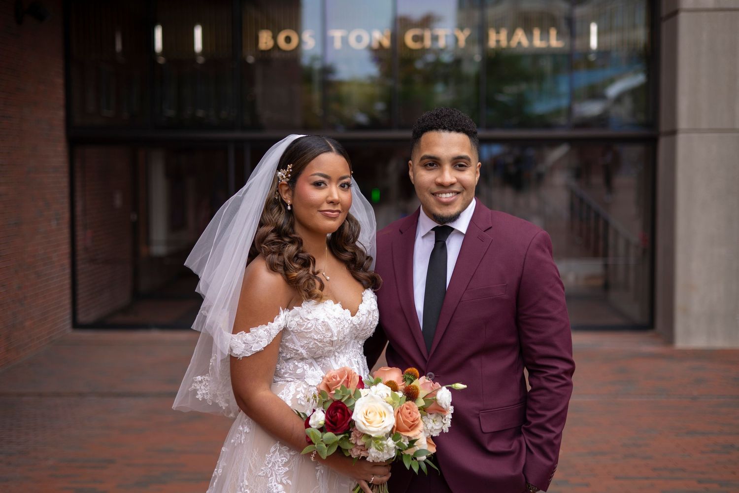 A wedding couple poses in front of Boston City Hall with brick architecture and signage in the background.