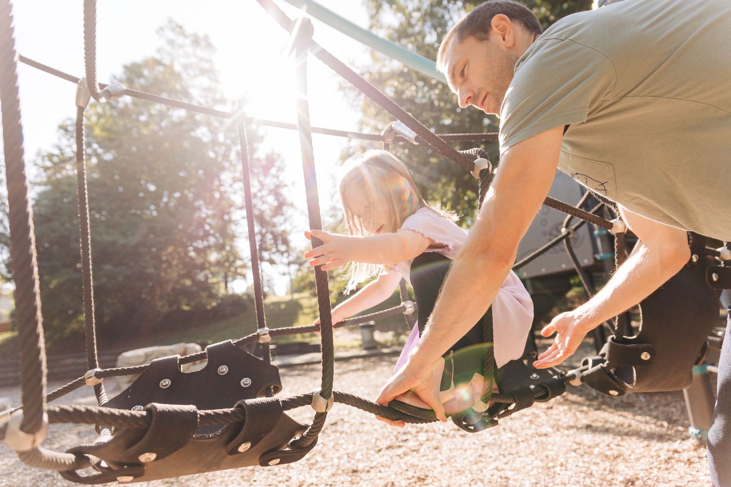 A dad helps a daughter cross a playground bridge with sunlight streaming through the trees.