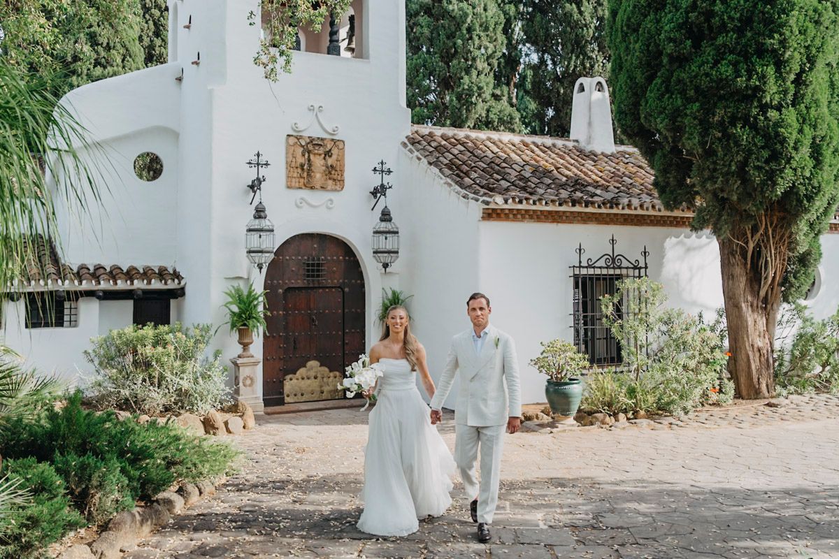 A couple in wedding attire walks along a stone path outside a Spanish-style white church with cypress trees.