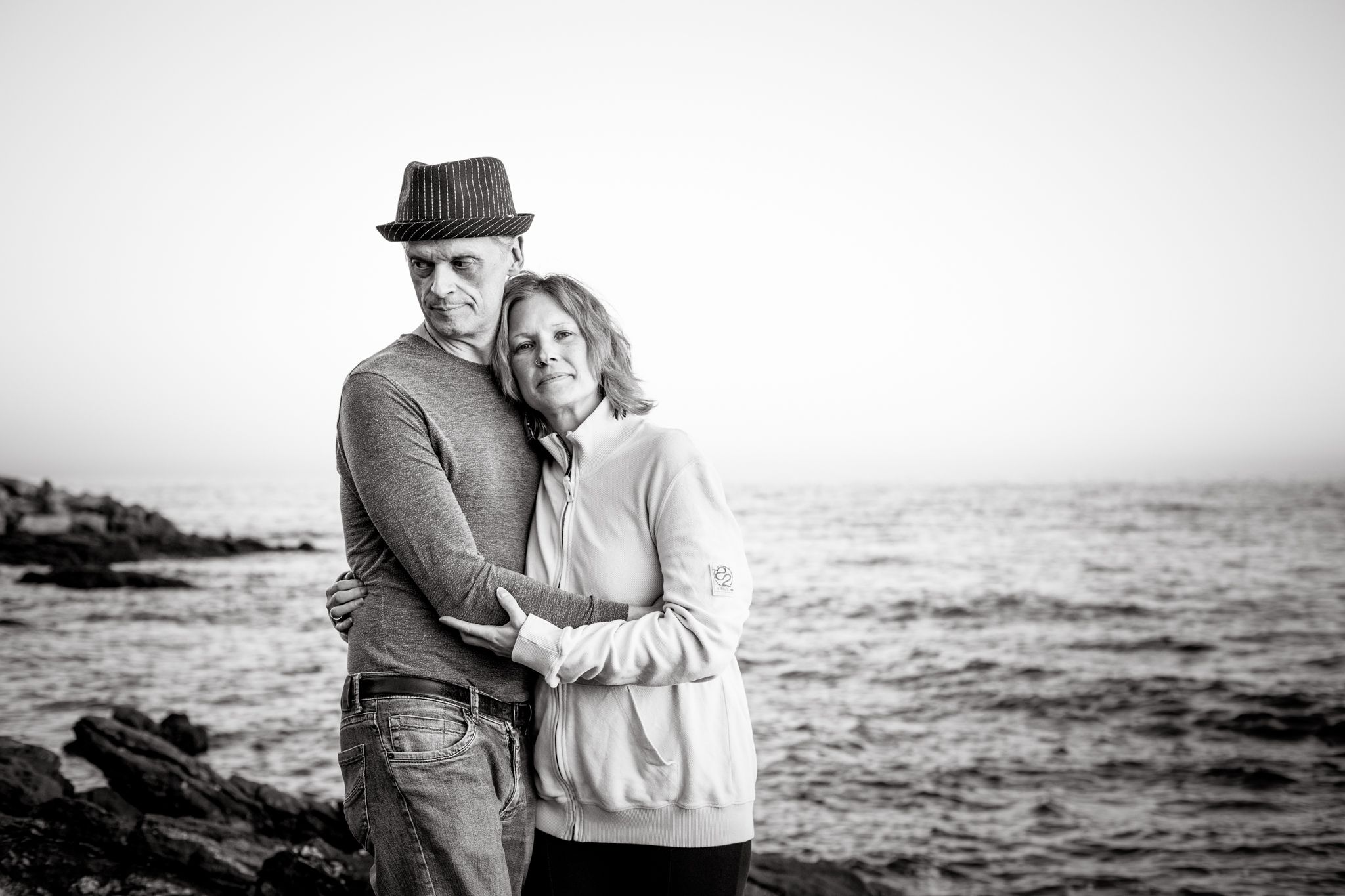 A black-and-white photo of a couple, hugging each other, with the sea behind, the woman looking to the camera.