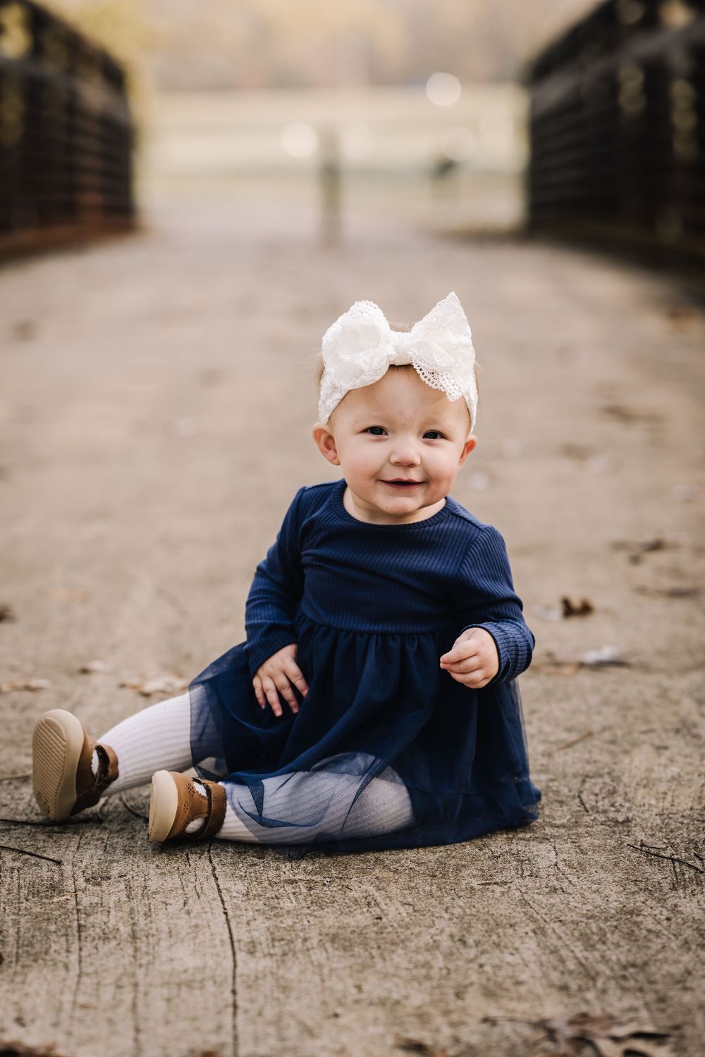 Baby in navy dress and white bow sitting on wooden bridge surface.