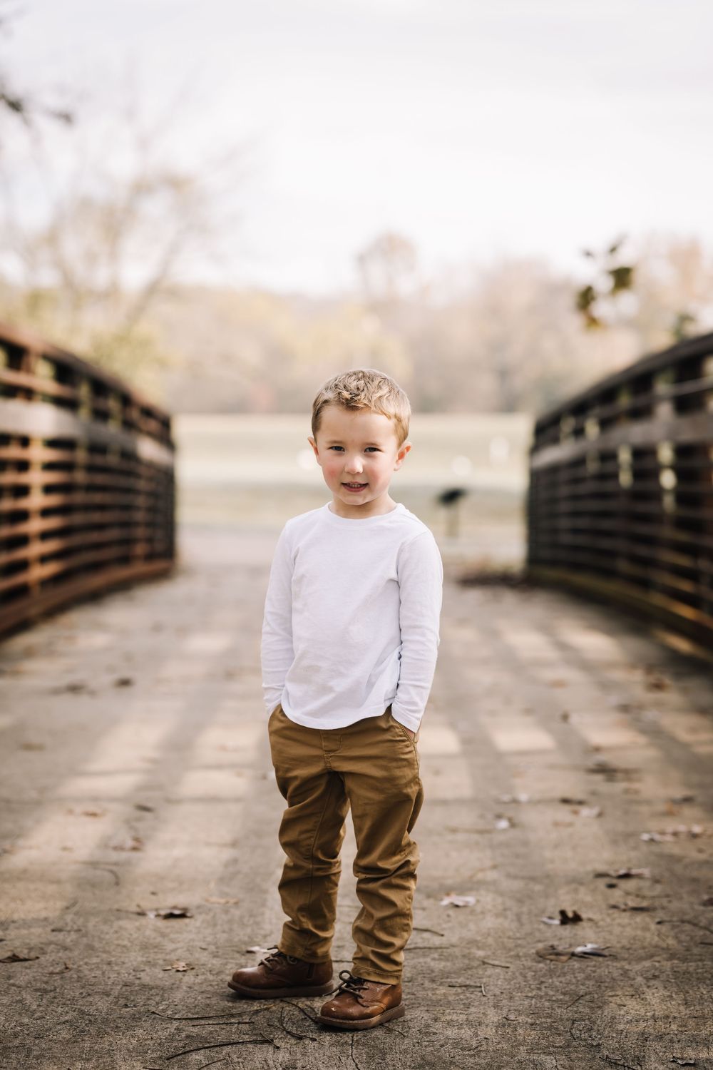 A young child stands alone on a wooden bridge wearing a white shirt and brown pants.