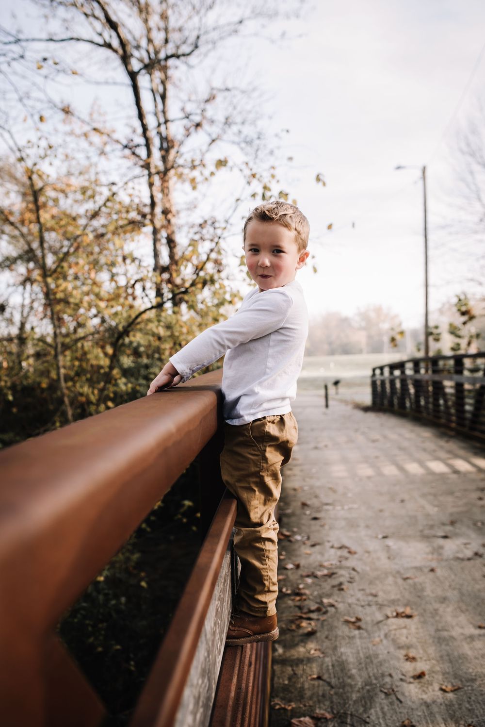 Young child in casual clothes leaning on wooden bridge railing during autumn.