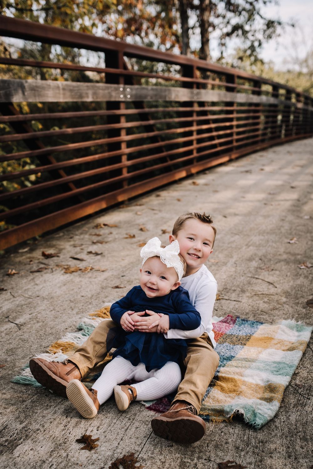 Two children sit together on a blanket on a wooden bridge with rustic metal railings on an autumn day.