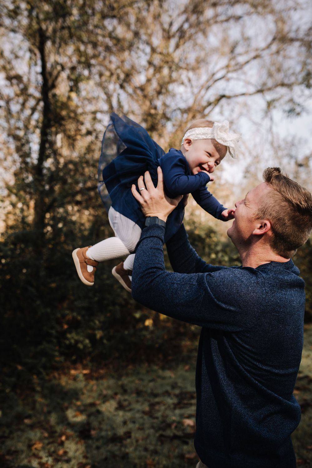 A tender moment captured outdoors as a parent lifts a small child wearing navy blue against an autumn backdrop.