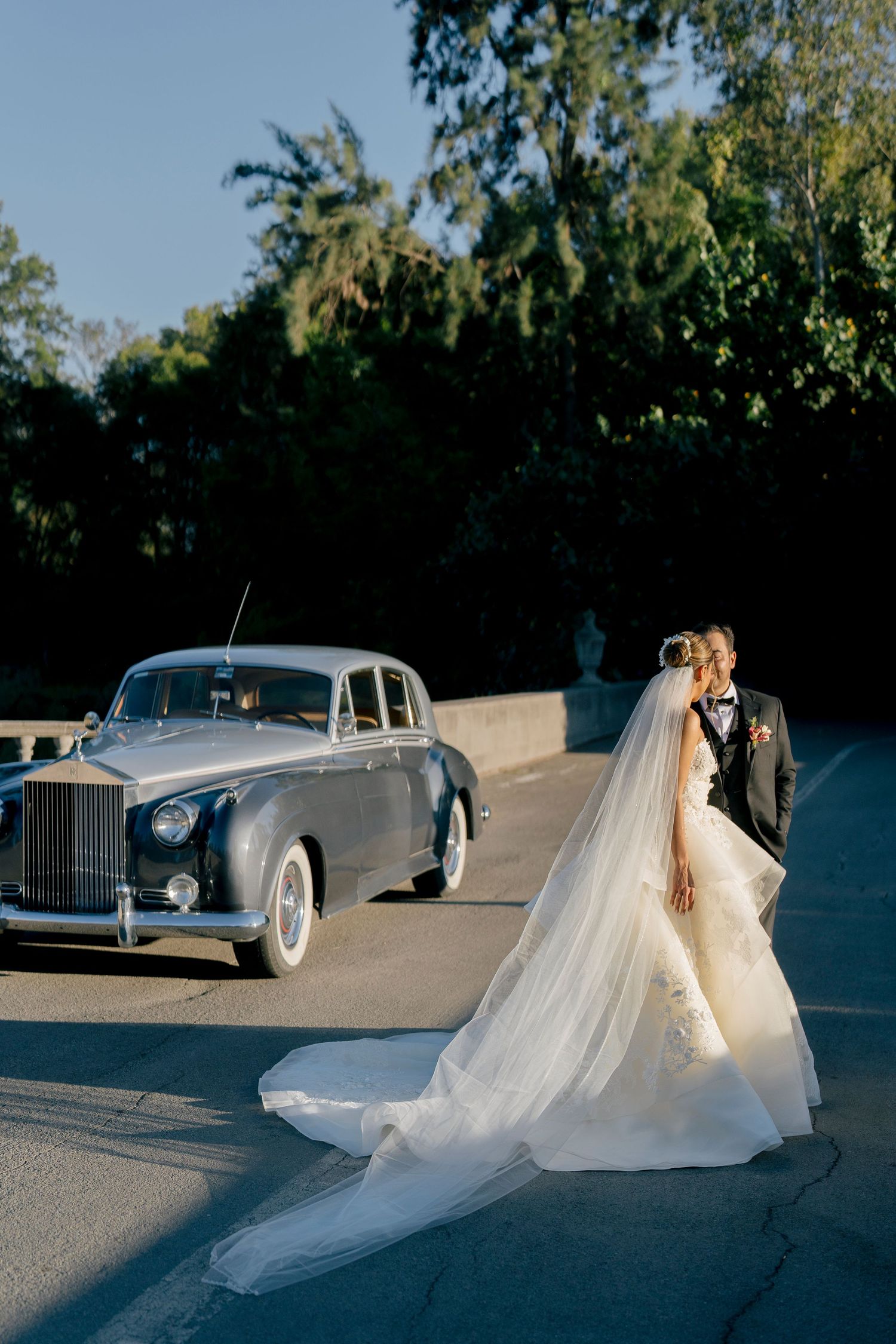 A classic silver Rolls Royce luxury car parked on a curved road with a flowing white wedding dress in the foreground.