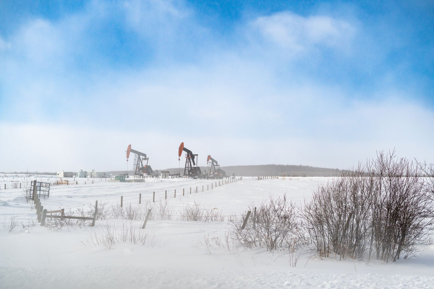 Three oil pump jacks operating in a vast snow-covered field under a cloudy winter sky.