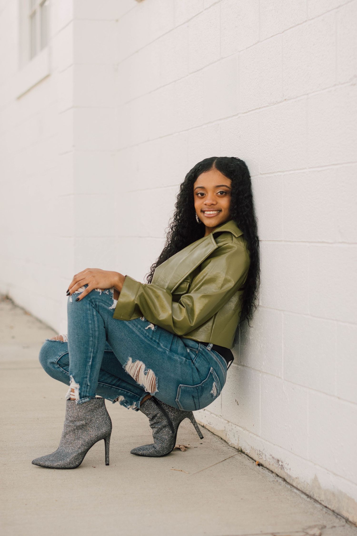 Stylish pose in olive jacket and distressed jeans against white wall.