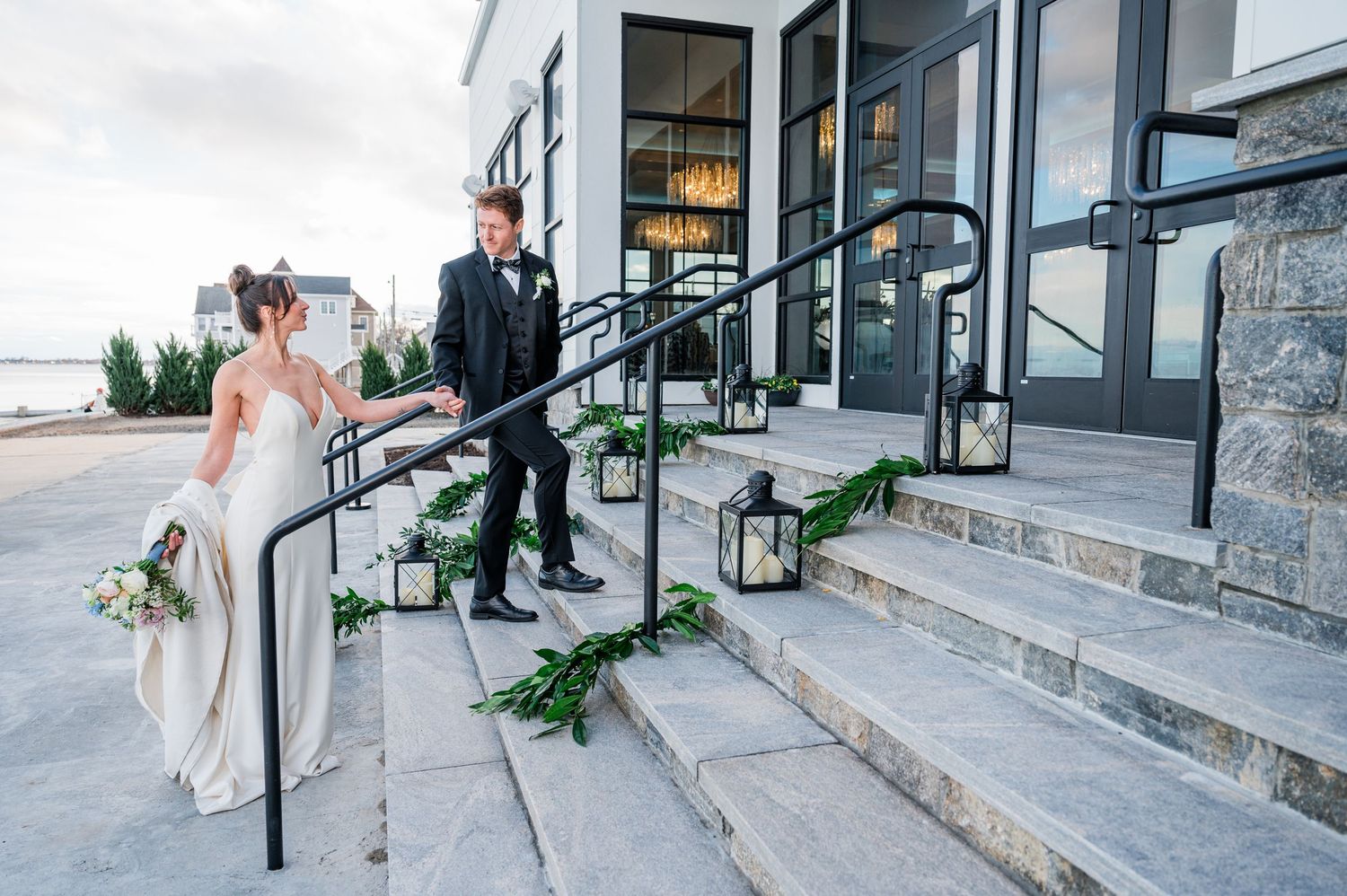 Elegant wedding couple embraces on stone steps decorated with lanterns and greenery at a rustic venue.