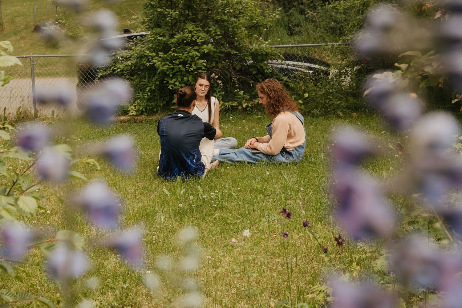 Three people sitting together in a flower-filled meadow, seen through soft purple wildflowers in the foreground.
