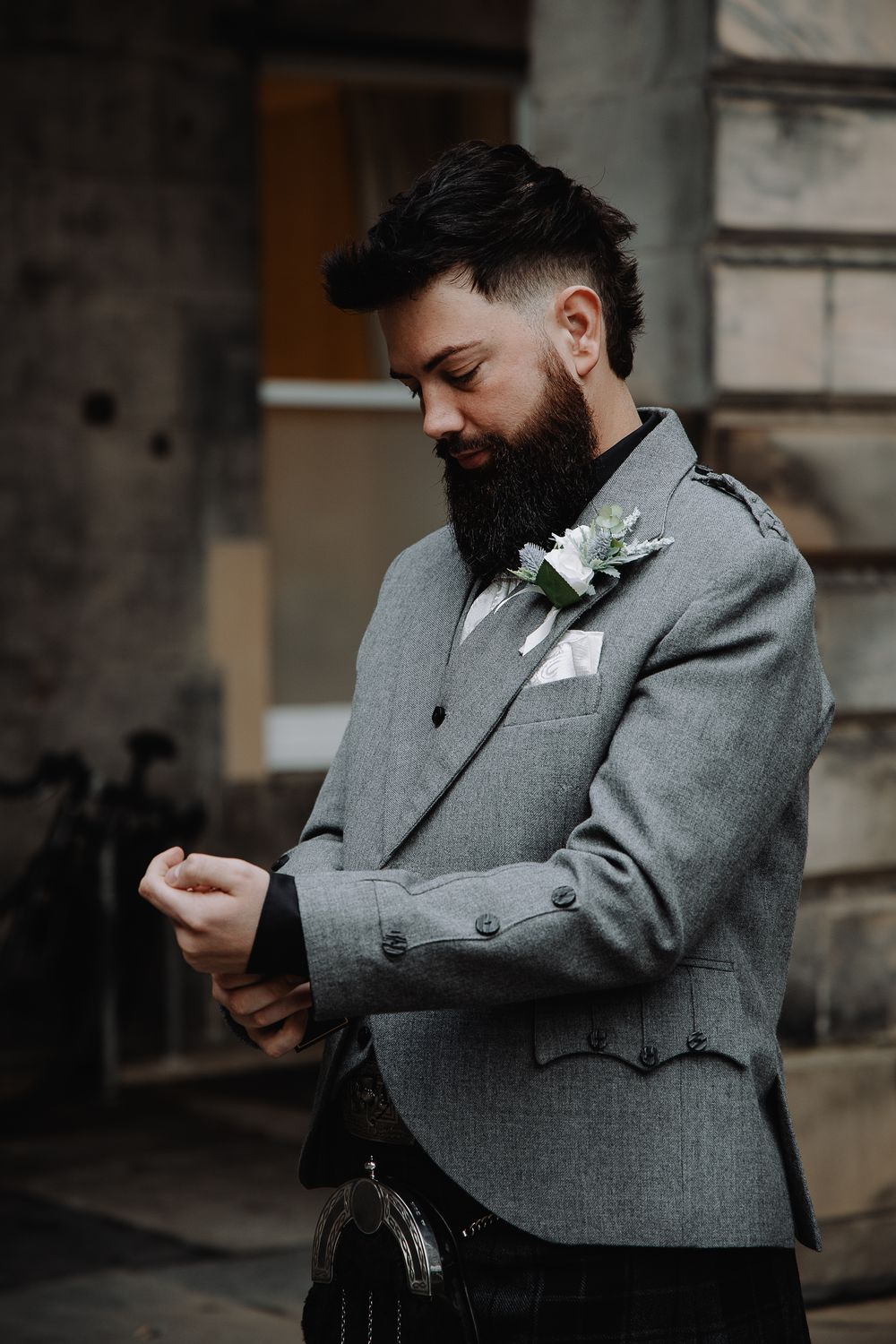 Bearded groom in gray Scottish kilt jacket with boutonniere adjusting cufflinks outdoors at wedding.